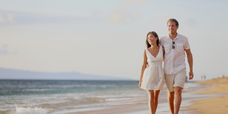 Pareja abrazada caminando por la orilla de la playa al atardecer, sonriendo y disfrutando de la vida.